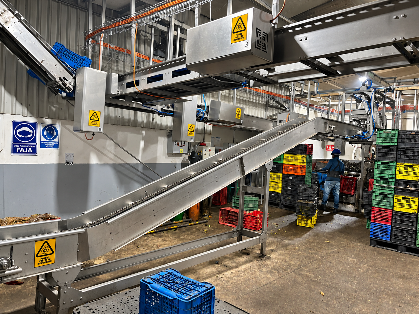 Línea de producción industrial en operación con transportadores de acero inoxidable, cajas de plástico y personal trabajando en planta.